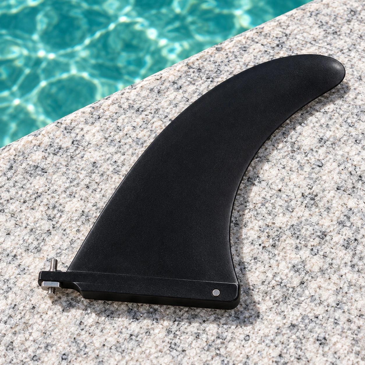 Black swept-back SUP center fin with its US-box mounting base on pale stone beside turquoise water